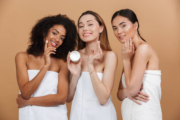 Beauty photo of three smilng multiracial women with different types of skin: caucasian, african american and asian girls, applying face cream together isolated over beige background - 写真・画像
