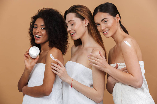 Beauty portrait of three young multiracial women with different types of skin: caucasian, african american and asian girls applying cream on body together isolated over beige background - Фото, зображення