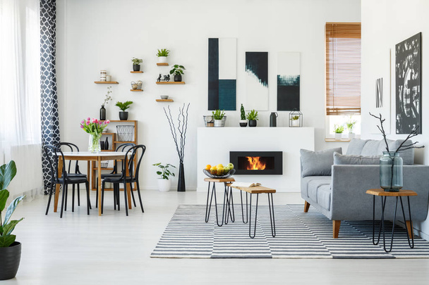 Black chairs at dining table in bright living room interior with grey sofa near fireplace. Real photo - Foto, Imagem