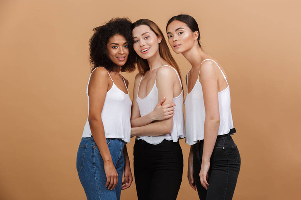 Image of three beautiful smiling women of different nation: caucasian, african american and asian girls in jeans standing together isolated over beige background - Foto, Imagem