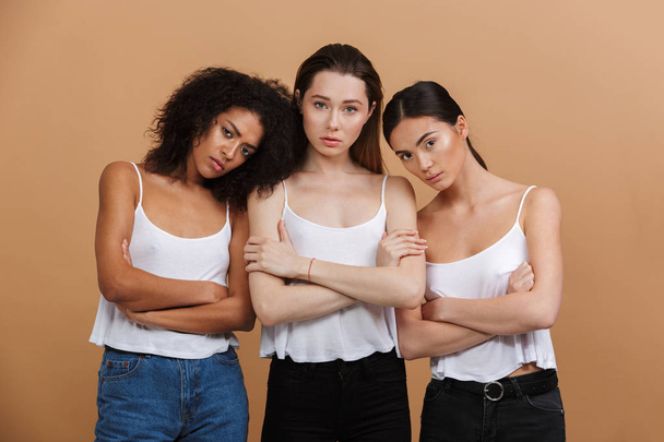Image of three serious interracial women: caucasian, african american and asian girls in jeans standing with arms crossed isolated over beige background - Фото, изображение
