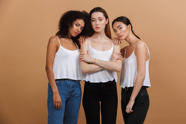Image of three beautiful international women: caucasian, african american and asian girls in casual clothing standing together isolated over beige background - Valokuva, kuva