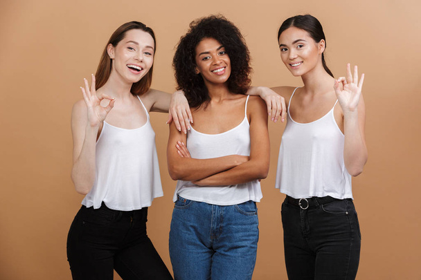 Three cheerful women posing together and showing peace gestures while looking at the camera over beige background - Foto, immagini