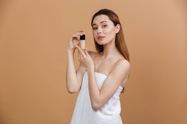 Beauty portrait of beautiful caucasian woman in white towel looking at camera and holding concealer bottle near face isolated over beige background - Foto, Imagem