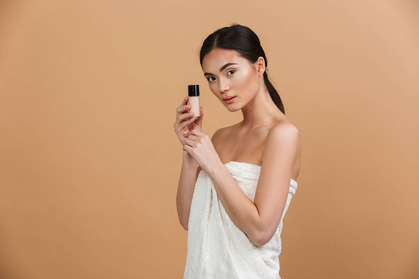Beauty portrait of brunette asian woman looking at camera and holding foundation bottle near face isolated over beige background - Foto, afbeelding