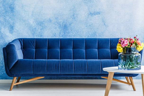 Comfortable navy blue plush sofa and colorful flowers in a vase on a table against ombre wall in a living room interior. Real photo. - Foto, afbeelding