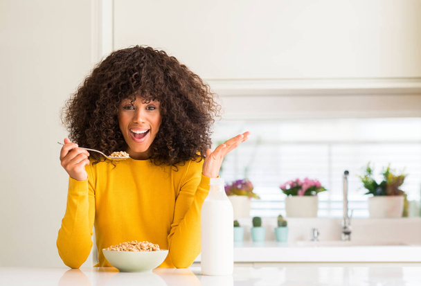 Mujer afroamericana comiendo cereales y leche en casa muy feliz y emocionada, expresión ganadora celebrando la victoria gritando con una gran sonrisa y levantando las manos
 - Foto, Imagen