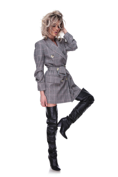 attractive woman wearing grey suit jacket and long leather boots posing with raised leg while fixing hair and looking down, full body picture - Photo, Image