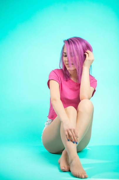 Portrait of bright young beautiful girl with pink colored hair smiling and looking at camera sitting on studio blue floor - Фото, изображение
