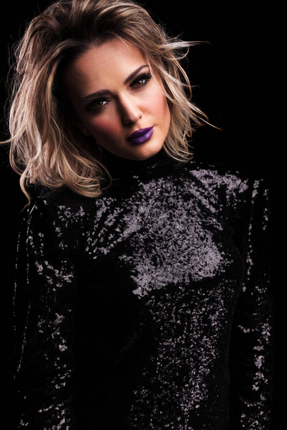 portrait of beautiful woman in black gleaming dress with purple lipstick leaning her head to side while standing on black background - Φωτογραφία, εικόνα