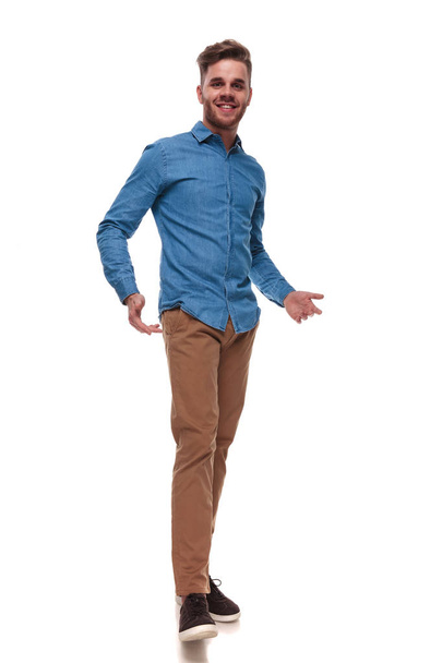 hombre casual feliz vistiendo una camisa azul de pie sobre fondo blanco y presentando, imagen de longitud completa
 - Foto, imagen