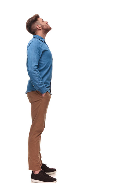 vista lateral del relajado hombre casual mirando algo mientras está de pie sobre fondo blanco, imagen de cuerpo entero
 - Foto, Imagen