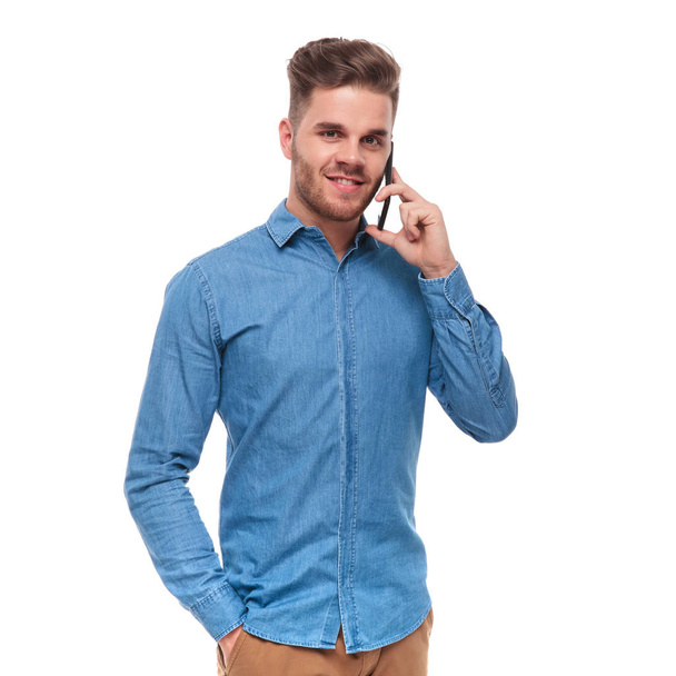 portrait of relaxed young casual man talking on the phone and standing on white background with hand in pocket - Zdjęcie, obraz