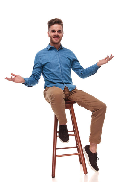 seated young casual man making a welcoming gesture with both hands on white background - Foto, imagen