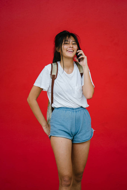 beautiful asian girl smiling talking on phone standing on studio red background - 写真・画像