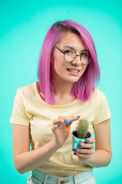 Beautiful woman with violet hair holding cactus in pot and touching thorns - Fotó, kép