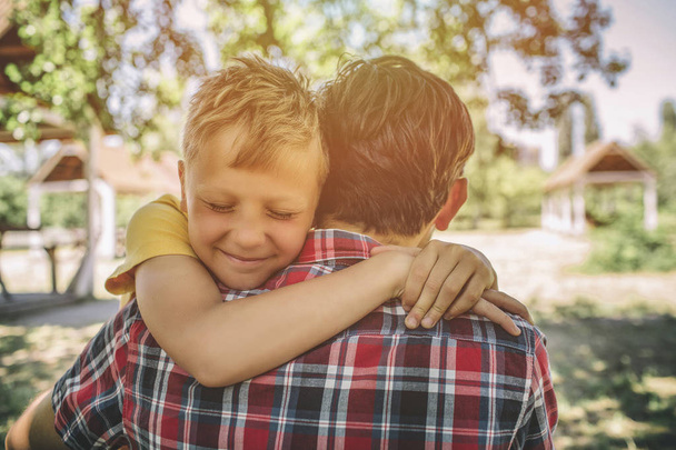 Cheerful and happy boy is hugging his father. Boy is keeping eyes closed and smiling. They are standing outside in park. - Foto, Imagen