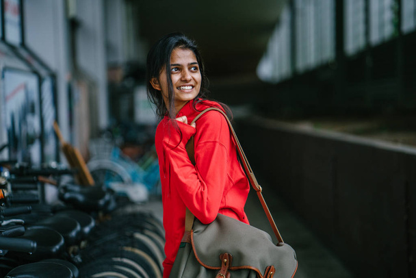 smiling indian girl with bag standing on dark background with parked bikes - Photo, image