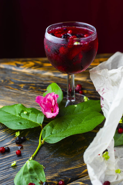 A glass full of red drink with berries, ice and a pink flower on crumpled wrapping paper beside which lie berries and a branch of green tropical leaves on a vintage wooden table. Copy space. - 写真・画像