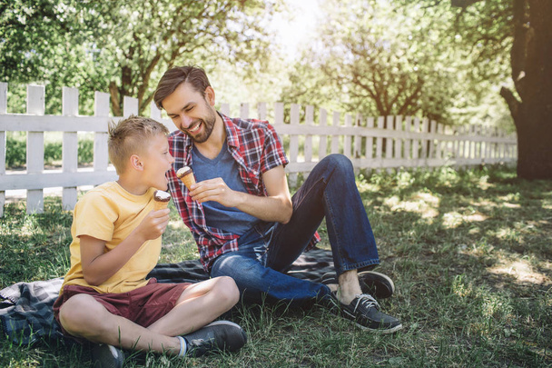 Nice picture of two guys sitting on blanket in park. They have ice creams in hands. Man is giving his son to try his ice cream. Child is opening mouth to bite a piece of ice cream. - Zdjęcie, obraz