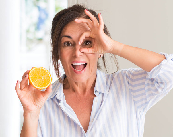 Μέσης ηλικίας γυναίκα που κρατώντας πορτοκαλί φρούτα με χαρούμενο πρόσωπο χαμογελά κάνει εντάξει σημάδι με το χέρι στο μάτι που αναζητούν μέσα από τα δάχτυλα - Φωτογραφία, εικόνα