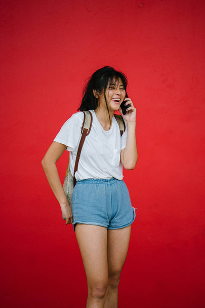 Krásná Asiatka s úsměvem mluvit na telefonu stojící na studio červené pozadí - Fotografie, Obrázek