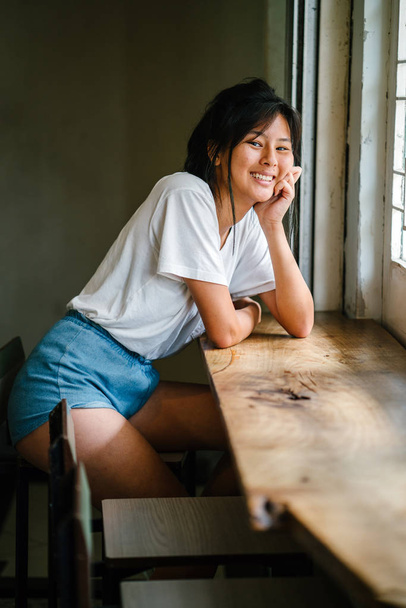 stylish asian girl sitting at wooden table near window - Foto, afbeelding