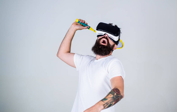 Man with beard in VR glasses play tennis, grey background. Guy with VR glasses play tennis with racket and ball. Hipster on busy face use modern technology for sport games. Virtual sport concept - Photo, image