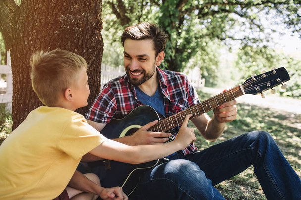 Small boy is leeaning to fathers guitar. He wants to play on it. Bearded guy is holding this guitar and smiling to son. He looks happy. - Fotografie, Obrázek