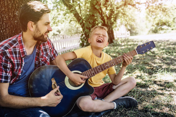 Joyful and satisfied young boy is holding guitar and playing on it. He is singing and keeping eyes closed. His father is helping him. He is proud of son. - Φωτογραφία, εικόνα