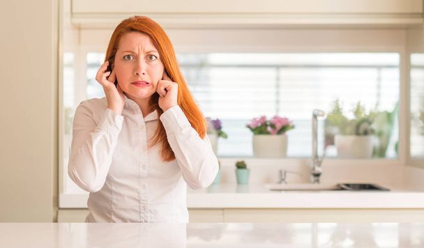 Рыжая женщина на кухне закрывает уши пальцами с раздраженным выражением для шума громкой музыки. Глухая концепция
. - Фото, изображение
