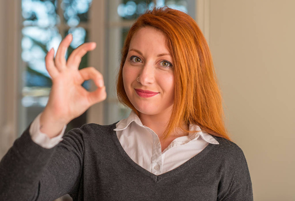 Κοκκινομάλλης γυναίκα στο σπίτι κάνει εντάξει πινακίδα με τα δάχτυλα, εξαιρετική σύμβολο - Φωτογραφία, εικόνα