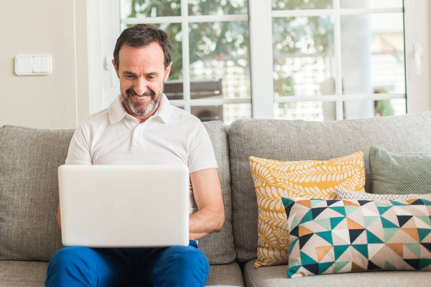Hombre de mediana edad utilizando el ordenador portátil en el sofá con una cara feliz de pie y sonriendo con una sonrisa confiada que muestra los dientes
 - Foto, imagen