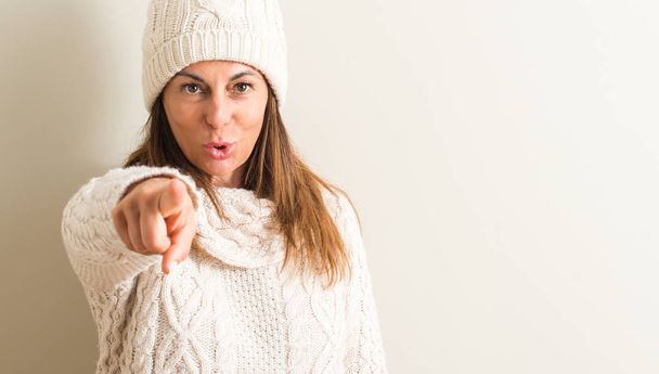Μέσης ηλικίας γυναίκα που φοράει καπάκι χειμώνα μαλλί, δείχνοντας με το δάχτυλο στη φωτογραφική μηχανή και σε εσάς, σημάδι χέρι, θετική και σίγουρη κίνηση από το μέτωπο - Φωτογραφία, εικόνα