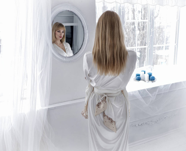 Bella giovane donna bionda in accappatoi bianchi in piedi accanto allo specchio in una stanza bianca
 - Foto, immagini