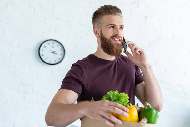 низький кут зору усміхненого бородатого чоловіка, який розмовляє по смартфону і тримає паперовий мішок зі свіжими овочами
  - Фото, зображення