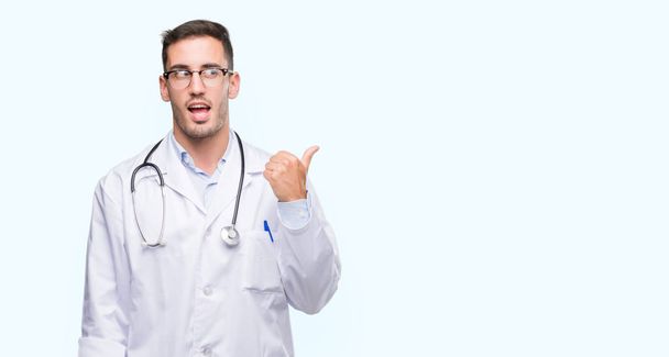 Handsome young doctor man smiling with happy face looking and pointing to the side with thumb up. - Photo, Image