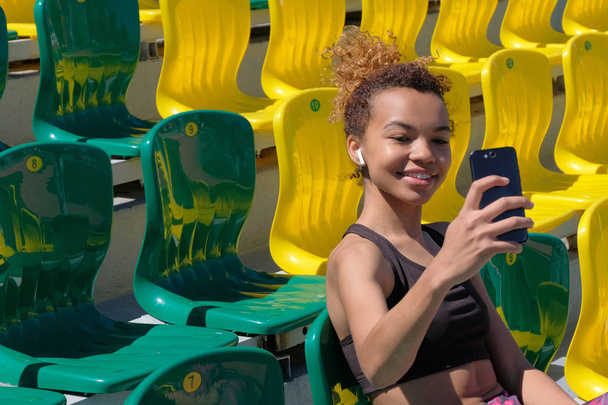 One beautiful, smiling, sexy African-American girl takes pictures of herself on the phone sitting on a green chair in the rows for the fans of the stadium. Sports lifestyle. - Photo, Image