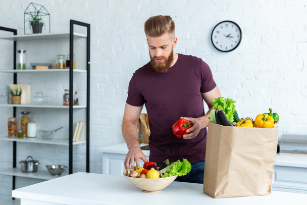 handsome bearded man putting fresh vegetables in bowl - Photo, Image