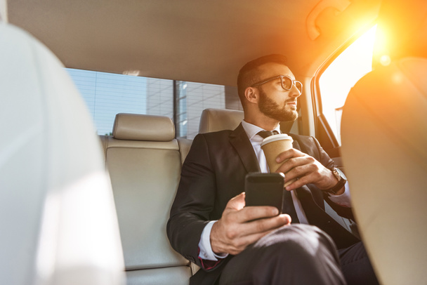 handsome businessman sitting in car with disposable coffee cup and smartphone - Photo, Image