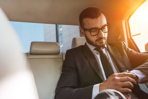 handsome businessman checking time on wristwatch in car during sunset - Foto, afbeelding