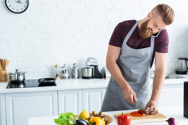 smiling bearded young man in apron cutting vegetables and talking by smartphone in kitchen - 写真・画像