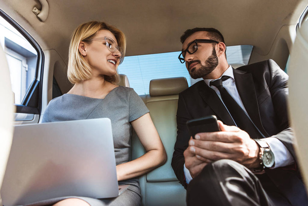 low angle view of handsome businessman and assistant working in car with laptop and smartphone - Photo, Image