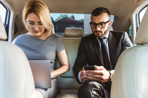 handsome businessman and assistant working in car with laptop and smartphone - Foto, afbeelding
