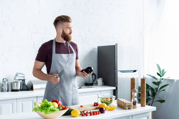 handsome bearded man in apron listening music in earphones and looking away while cooking in kitchen  - Photo, Image