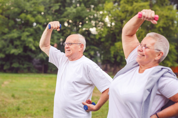 Uśmiechnięty starszy mężczyzna i kobieta są na świeżym powietrzu w parku ćwiczenia z hantlami i wspólna zabawa - Zdjęcie, obraz
