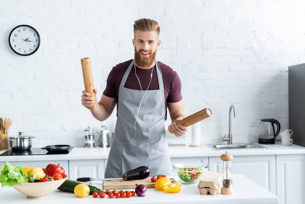 красивый бородатый мужчина в фартуке слушает музыку в наушниках и улыбается в камеру во время приготовления пищи на кухне
 - Фото, изображение