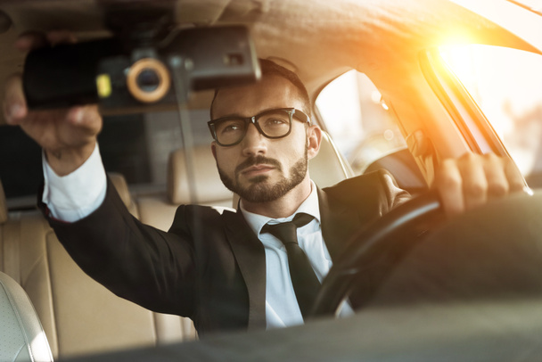 handsome driver in suit driving car and fixing mirror during sunset - Photo, Image
