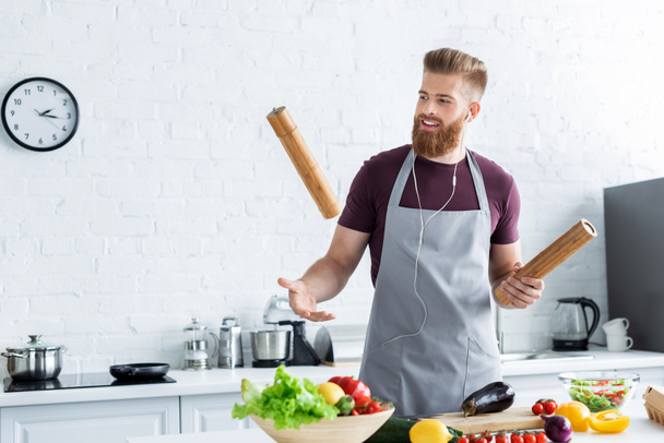 handsome smiling bearded man in apron listening music in earphones while cooking in kitchen - Photo, Image