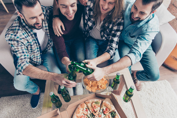 Cropped top view of four bearded, stylish, professional, successful, confident, attractive guys in casual, denim outfit clinking bottles with lager, having snacks on the table, group embracing - Фото, изображение